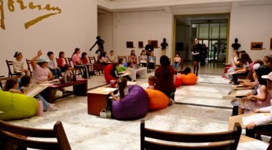 People, including children, sit in a circle of colorful bean bags and chairs in a gallery as a presenter talks at the front.