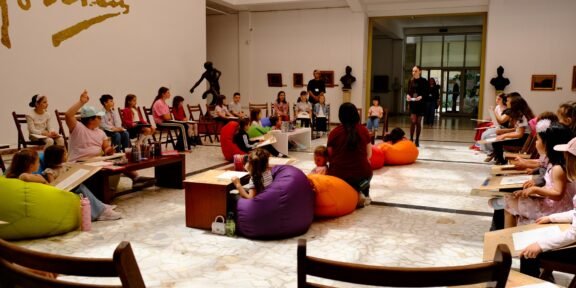 People, including children, sit in a circle of colorful bean bags and chairs in a gallery as a presenter talks at the front.