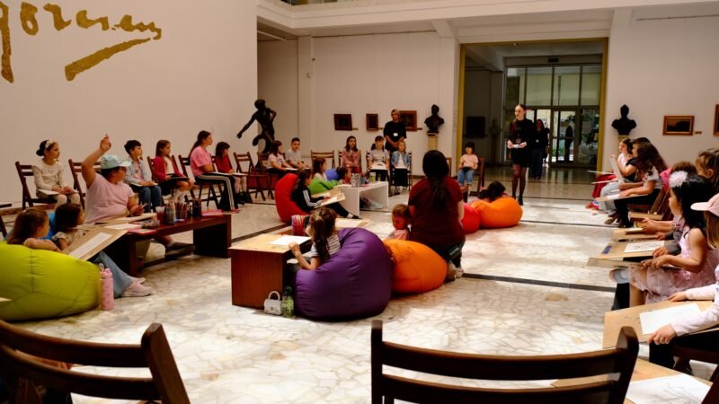 People, including children, sit in a circle of colorful bean bags and chairs in a gallery as a presenter talks at the front.