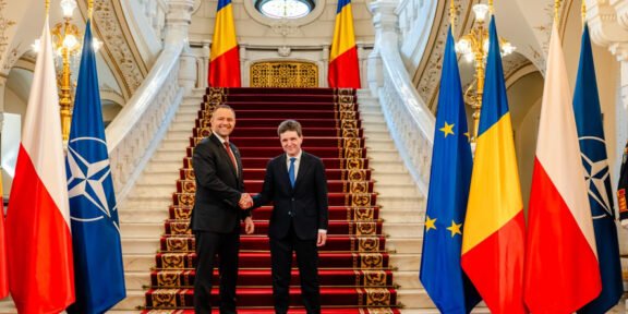 Two men in suits shake hands at a grand staircase, flanked by flags including Poland, NATO, EU, and Romania