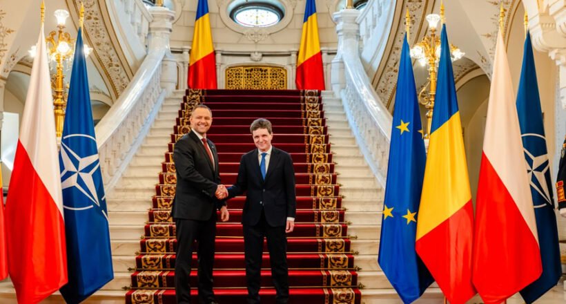 Two men in suits shake hands at a grand staircase, flanked by flags including Poland, NATO, EU, and Romania