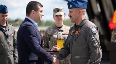 Civilian man in a dark suit shakes hands with a uniformed military officer during an award ceremony, with teammates in uniform in the background.
