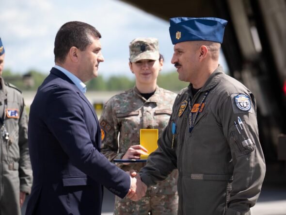 Civilian man in a dark suit shakes hands with a uniformed military officer during an award ceremony, with teammates in uniform in the background.