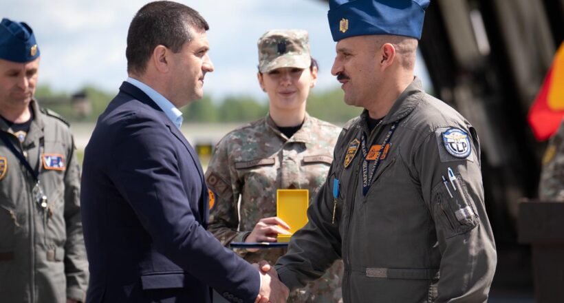 Civilian man in a dark suit shakes hands with a uniformed military officer during an award ceremony, with teammates in uniform in the background.
