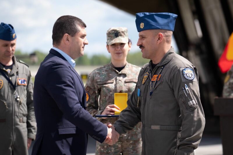 Civilian man in a dark suit shakes hands with a uniformed military officer during an award ceremony, with teammates in uniform in the background.