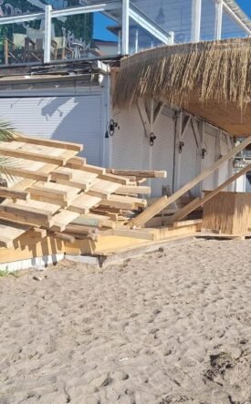 Stacks of wooden planks on a sandy beach beside a thatched-roof shed, with palm plants and a blue ocean in the background.