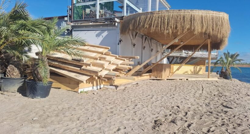 Stacks of wooden planks on a sandy beach beside a thatched-roof shed, with palm plants and a blue ocean in the background.