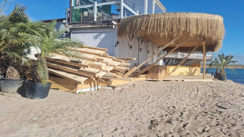 Stacks of wooden planks on a sandy beach beside a thatched-roof shed, with palm plants and a blue ocean in the background.