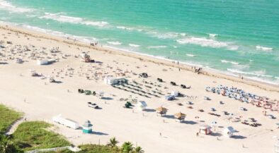 Aerial view of a sunny beach with turquoise water, rows of umbrellas and lounge chairs on the sand, and a palm‑lined promenade in the foreground.