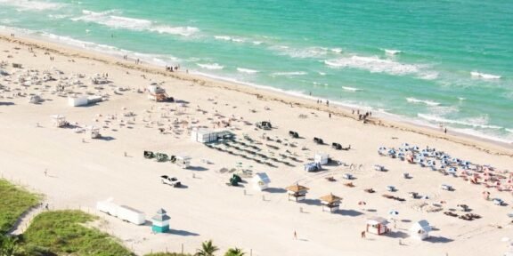 Aerial view of a sunny beach with turquoise water, rows of umbrellas and lounge chairs on the sand, and a palm‑lined promenade in the foreground.