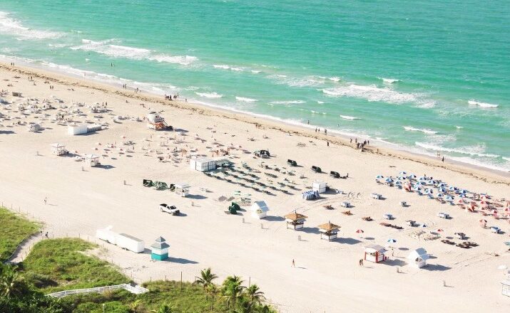 Aerial view of a sunny beach with turquoise water, rows of umbrellas and lounge chairs on the sand, and a palm‑lined promenade in the foreground.