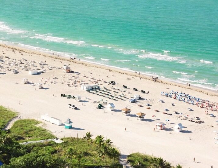 Aerial view of a sunny beach with turquoise water, rows of umbrellas and lounge chairs on the sand, and a palm‑lined promenade in the foreground.