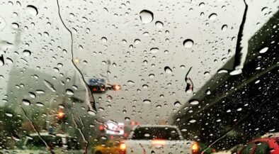 Rain-speckled windshield view of a congested highway with blurred tail lights in gray weather behind cars ahead on a bridge or overpass