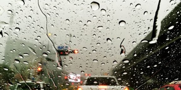 Rain-speckled windshield view of a congested highway with blurred tail lights in gray weather behind cars ahead on a bridge or overpass