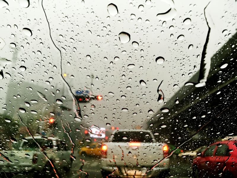 Rain-speckled windshield view of a congested highway with blurred tail lights in gray weather behind cars ahead on a bridge or overpass