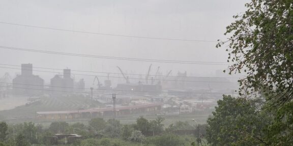 Industrial port and refinery structures with cranes visible through a hazy atmosphere, seen behind a curved railway track and greenery in the foreground.