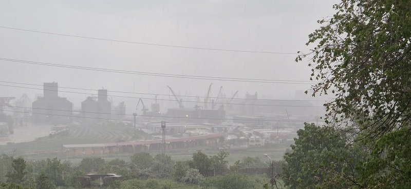 Industrial port and refinery structures with cranes visible through a hazy atmosphere, seen behind a curved railway track and greenery in the foreground.