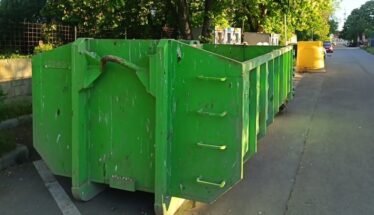 Green dumpsters lined up along a city street, curbside with trees in the background and a yellow barrier in the distance.