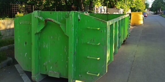Green dumpsters lined up along a city street, curbside with trees in the background and a yellow barrier in the distance.