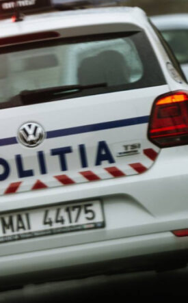 Rear view of a white Romanian police car with 'POLITIA' on the trunk and red emergency lights flashing on the roof, driving on a street.