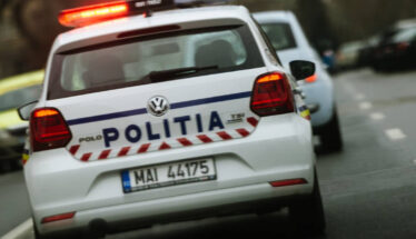 Rear view of a white Romanian police car with 'POLITIA' on the trunk and red emergency lights flashing on the roof, driving on a street.