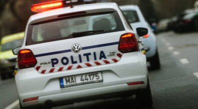Rear view of a white Romanian police car with 'POLITIA' on the trunk and red emergency lights flashing on the roof, driving on a street.