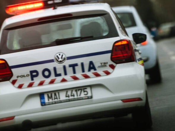 Rear view of a white Romanian police car with 'POLITIA' on the trunk and red emergency lights flashing on the roof, driving on a street.