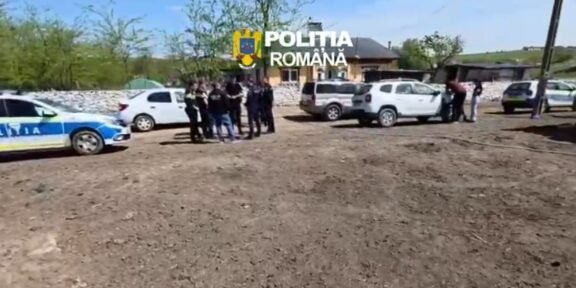 Group of police officers and civilians gathered near parked cars on a dirt lot in a rural area; Romanian police car visible.