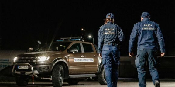 Two border police officers in uniform walk away from a patrol pickup at night, a 'Politia de Frontiera' vehicle nearby.