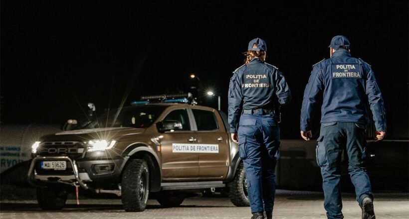 Two border police officers in uniform walk away from a patrol pickup at night, a 'Politia de Frontiera' vehicle nearby.