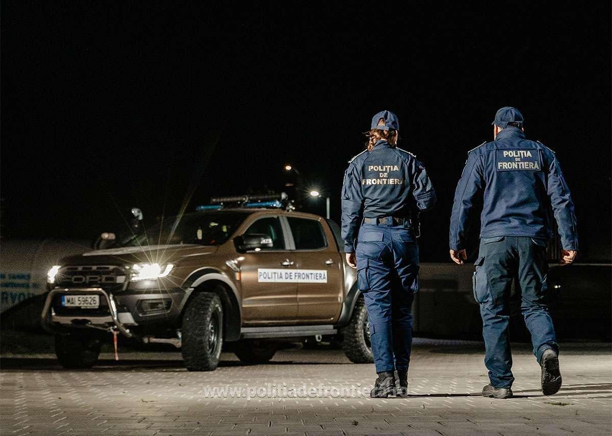 Two border police officers in uniform walk away from a patrol pickup at night, a 'Politia de Frontiera' vehicle nearby.