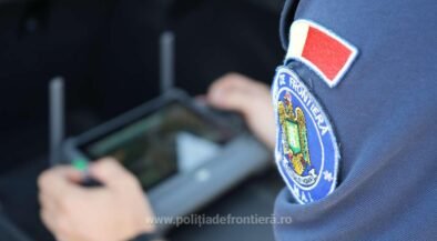 Close-up of a Romanian border police patch on a blue uniform; an officer uses a handheld tablet in the blurred background.