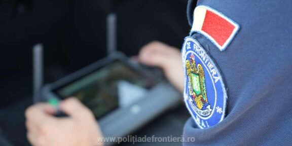 Close-up of a Romanian border police patch on a blue uniform; an officer uses a handheld tablet in the blurred background.