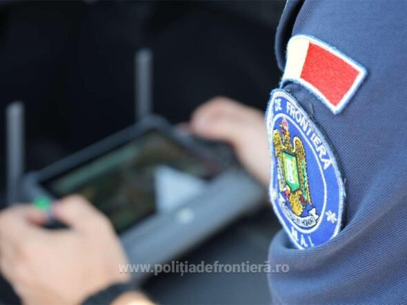 Close-up of a Romanian border police patch on a blue uniform; an officer uses a handheld tablet in the blurred background.