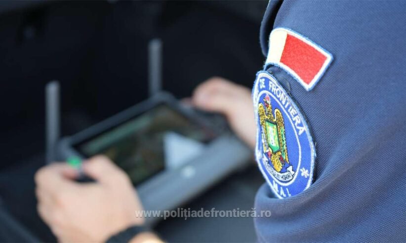 Close-up of a Romanian border police patch on a blue uniform; an officer uses a handheld tablet in the blurred background.