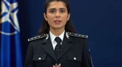 Female police officer in uniform delivering a speech, hands clasped, with a blue NATO-style flag in the background.