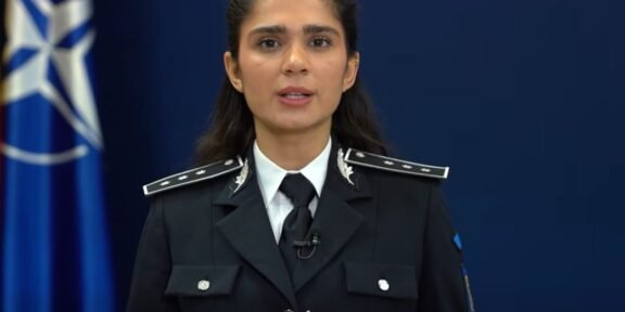 Female police officer in uniform delivering a speech, hands clasped, with a blue NATO-style flag in the background.
