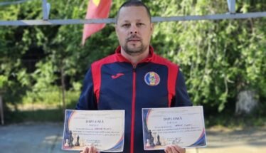Man in a blue-red sports jacket holds two diplomas outdoors, with greenery in the background and colorful flags above him.