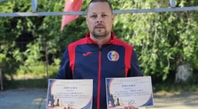 Man in a blue-red sports jacket holds two diplomas outdoors, with greenery in the background and colorful flags above him.