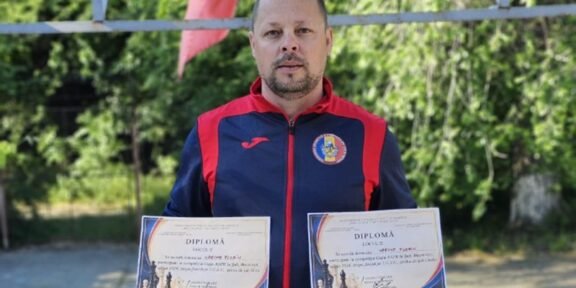 Man in a blue-red sports jacket holds two diplomas outdoors, with greenery in the background and colorful flags above him.