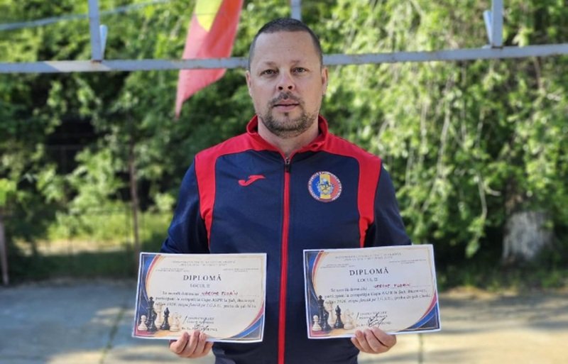 Man in a blue-red sports jacket holds two diplomas outdoors, with greenery in the background and colorful flags above him.