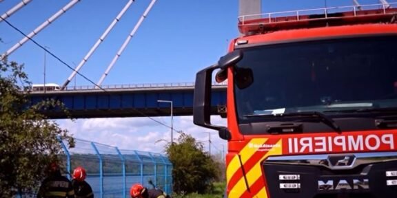 Firefighters in protective gear set up equipment on an orange mat beside a red fire truck under a cable-stayed bridge.