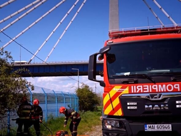 Firefighters in protective gear set up equipment on an orange mat beside a red fire truck under a cable-stayed bridge.