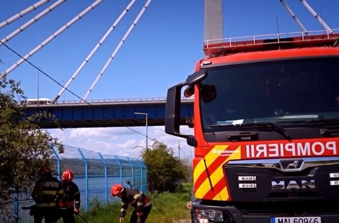 Firefighters in protective gear set up equipment on an orange mat beside a red fire truck under a cable-stayed bridge.