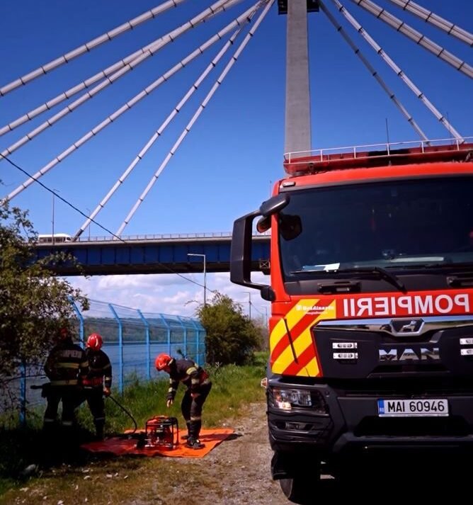 Firefighters in protective gear set up equipment on an orange mat beside a red fire truck under a cable-stayed bridge.