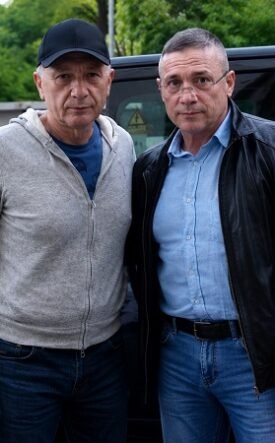 Two men pose together in front of a black van in a parking lot, dressed casually in jeans and jackets.