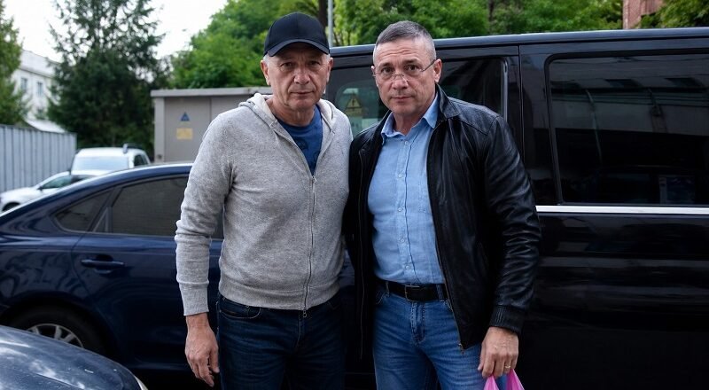 Two men pose together in front of a black van in a parking lot, dressed casually in jeans and jackets.