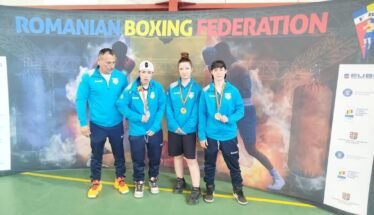 Coach and three young boxers in blue jackets pose with medals in front of a Romanian Boxing Federation banner indoors.