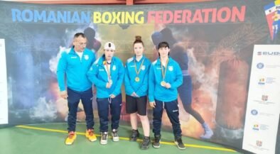 Coach and three young boxers in blue jackets pose with medals in front of a Romanian Boxing Federation banner indoors.
