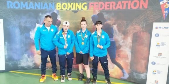 Coach and three young boxers in blue jackets pose with medals in front of a Romanian Boxing Federation banner indoors.